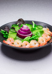 Green cucumber and arugula. Salad with cabbage leaves and golden shrimps. Black dish. Dark background. White table. Concept for a tasty and healthy meal. Crimean onion.