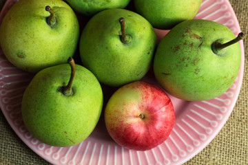 Green yellow organic pears and one small red apple, different not like others, on delicate pink plate, on a coarse retro cloth canvas. Dark atmospheric photo of fruits, autumn harvest. Top view 
