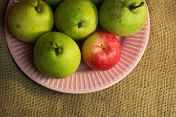 Green yellow organic pears and one small red apple, different not like others, on delicate pink plate, on a coarse retro cloth canvas. Dark atmospheric photo of fruits, autumn harvest. Top view 
