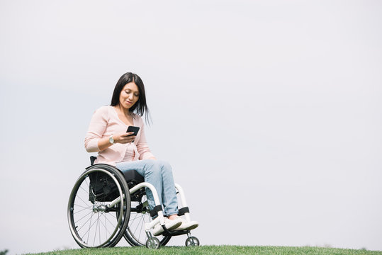 smiling disabled woman in wheelchair using smartphone while resting in park - Powered by Adobe