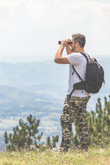 Traveler with backpack standing on the top of the mountain and using binoculars