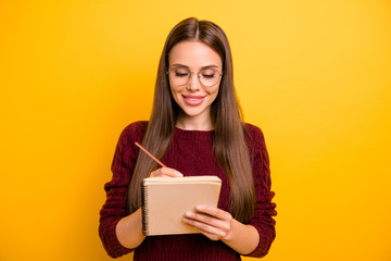 Portrait of charming girl holding pen and notebook writing check plan to do list wearing maroon jumper isolated over yellow background