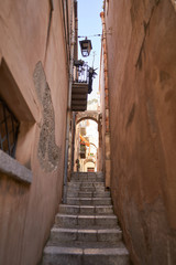 Fototapeta premium Thin or narrow small street in sicilian city Cefalu with marble stone stairs hanging lantern and balcony. Typical example of historic and traditional mediterranean architecture taken in hot sunny day