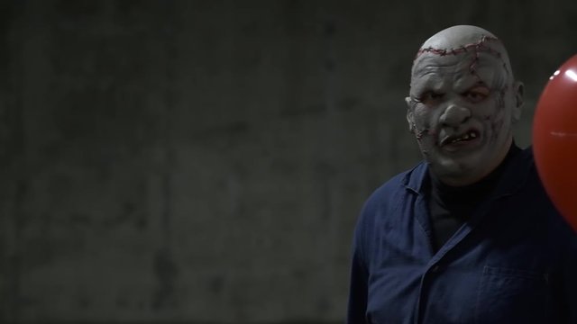 Creepy man in leather mask with red scars, in blue jumpsuit, with baseball bat and red ball is standing against wall, slow motion, monster raises bat, looks menacingly at camera