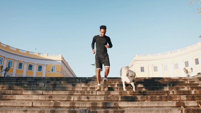 Young black man running with his white labrador dog down the stairs in old city center during morning, slow motion