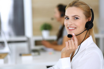 Blonde business woman using headset for communication and consulting people at customer service office. Call center. Group of operators at work at the background - obrazy, fototapety, plakaty