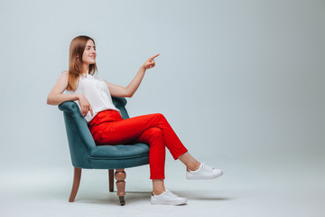 girl in red pants sits in a chair and points at a light gray background. copy space