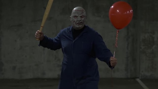 Frightening, scary man in leather mask, with bat and red ball in his hand threatensly bows at viewer, angrily looks at camera, approaches in slow motion, deaf concrete basement, maniac attacks