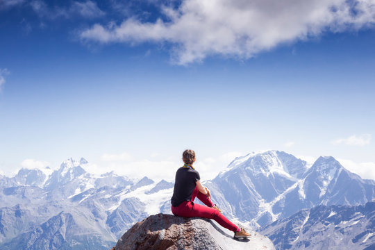 a young active girl sits on the edge of a rock, look at the Caucasus mountains. girl on backdrop of mountains sits its back