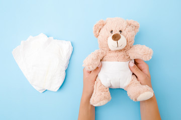Woman hands holding brown teddy bear with white diaper on pastel blue background. Closeup.