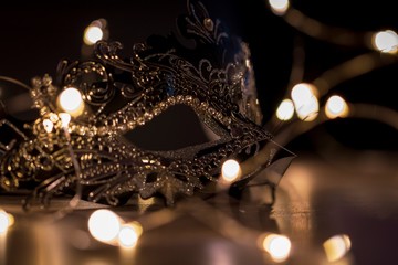 A portrait of a venetian mask on a wooden table surrounded by fairy lights. The mask is to be worn to hide someones identity on a masked ball.