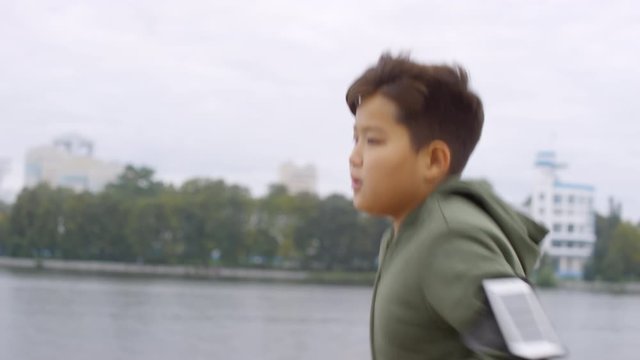 Side view of young Asian boy with smartphone in armband running along urban riverside in the morning