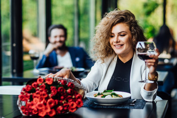 Girl is sitting at a table in the restaurant and drink wine, enjoy bouquet of red roses flower and waiting a date.