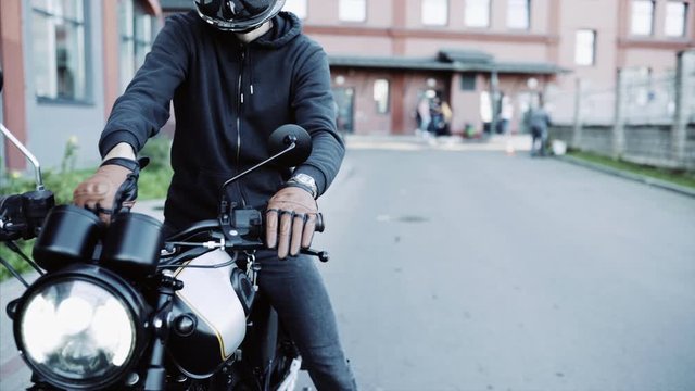Biker in leather gloves and black hoodie starts motorcycle and drives. Motorcyclist and vintage motorbike at parking space, modern, trendy safety helmet. Side view urban lifestyle scene