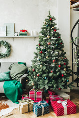 Christmas / New Year composition. Winter holidays concept. Beautiful scandinavian living room decorated with festive christmas fir tree with red, gold and green toys and big gift boxes.