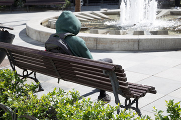 Mysterious and suspicious young man wearing winter hooded jacket in the middle of summer, sitting on the wooden bench alone, with a backpack on his back.