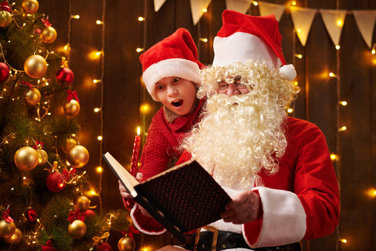Santa Claus and santa helper boy reading book, sitting indoor near decorated xmas tree with lights - Merry Christmas and Happy Holidays!