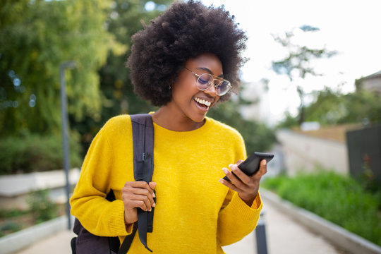smiling young black woman with glasses and bag looking at cellphone outdoors