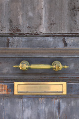 Brass handle and shiny mail slot on a vintage grey door with weathered cracked paint.