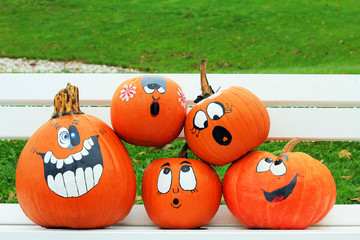 Orange pumpkins with funny drawn ghost faces are on the bench as decoration for outdoor autumn holiday in the park. Funny vegetables. Harvest and halloween concept.