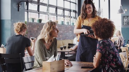 Two Female Friends Sitting At Table In Coffee Shop Being Served By Waitress