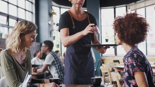 Two Female Friends Sitting At Table In Coffee Shop Being Served By Waiter