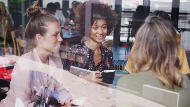 Four Young Female Friends Meeting Sitting At Table In Coffee Shop And Talking Viewed Through Window