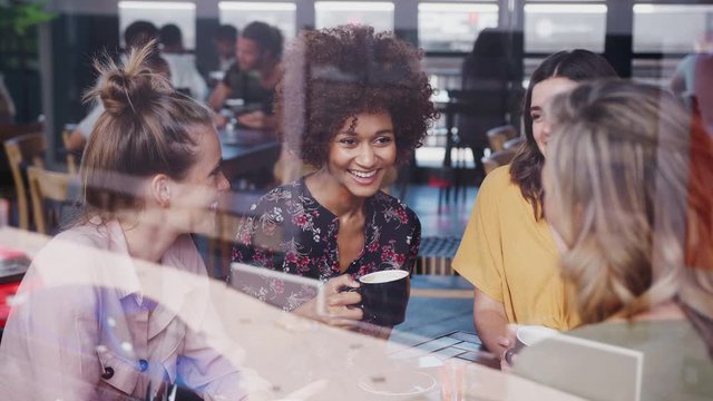 Four Young Female Friends Meeting Sitting At Table In Coffee Shop And Talking Viewed Through Window