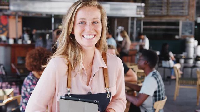Portrait Of Waitress Holding Digital Tablet In Busy Bar Restaurant Smiling At Camera