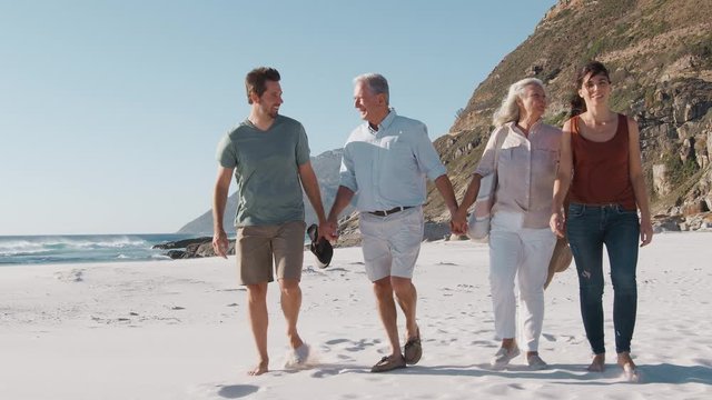 Senior Parents With Adult Offspring Walking Along Sandy Beach On Summer Vacation