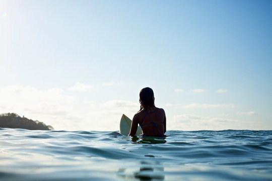 Silhouette female surfer straddling surfboard, waiting in sunny blue ocean
