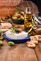 Greek cheese feta with thyme and olives, selective focus. Bread and young olives branch on olive board over old wood background. Overhead view.