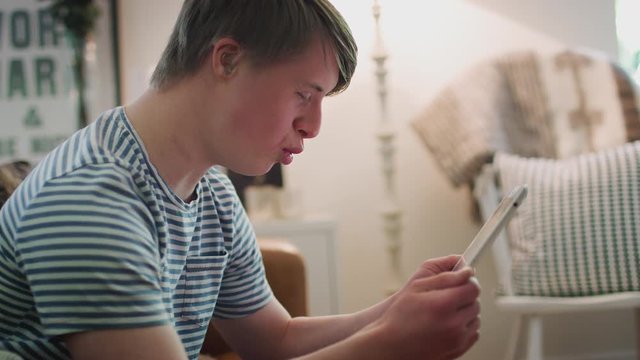 Young Downs Syndrome Man Sitting On Sofa Watching Digital Tablet At Home