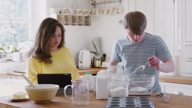 Young Downs Syndrome Couple Following Recipe On Digital Tablet To Bake Cake In Kitchen At Home