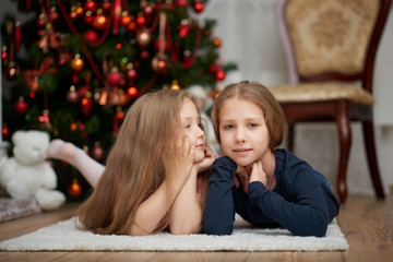 Two young sisters laying on white carpet. The little one is looking at the big one, which is looking at camera. Decorated Christmas tree background.