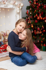 Fototapeta na wymiar Two young sisters sitting and hugging on white carpet. Fireplace and decorated Christmas tree background.