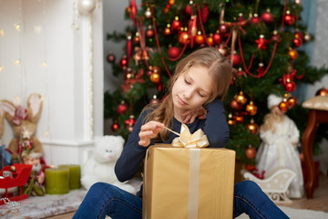 Obraz na płótnie Canvas A young girl wearing a blue long sleeve shirt and jeans sitting on white carpet. Fireplace filled with toys and decorated Christmas tree background.