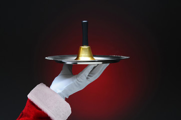 Closeup of Santa Claus hand holding a bell on a silver tray. Horizontal format on a light to dark red background.