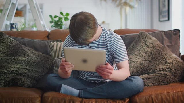 Young Downs Syndrome Man Sitting On Sofa Watching Digital Tablet At Home