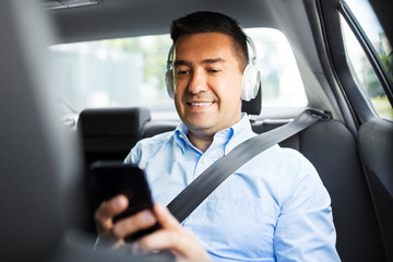 transport, business and technology concept - male passenger or businessman with wireless headphones using smartphone on back seat of taxi car
