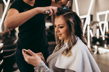 Beautiful woman with long hair at the beauty salon using smart phone and choosing hairstyle while getting a hair blowing. Hair salon styling concept.