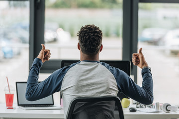 back view of african american programmer showing thumbs up while sitting at workplace near window