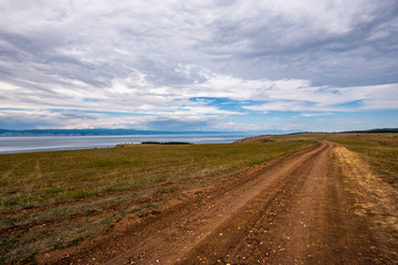 Fototapeta na wymiar A dirt brown road stretches into the distance along the shore of a lake in cloudy weather. Behind the lake are mountains, the sky is overcast.