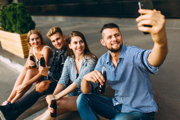 A group of cheerful friends, guys and girls walk around the city taking selfies on a smartphone, having fun and drinking drinks