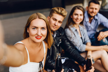 A group of cheerful friends, guys and girls walk around the city taking selfies on a smartphone, having fun and drinking drinks