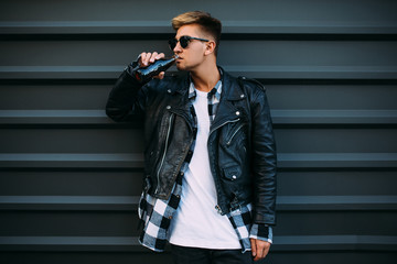A handsome man in sunglasses, in a leather black jacket posing and drinking a drink near a black wall