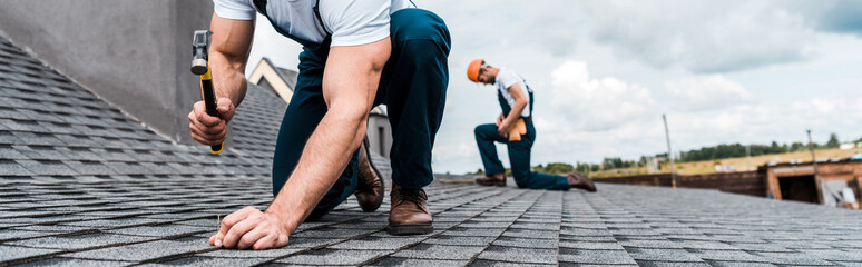 panoramic shot of handyman holding hammer while repairing roof near coworker