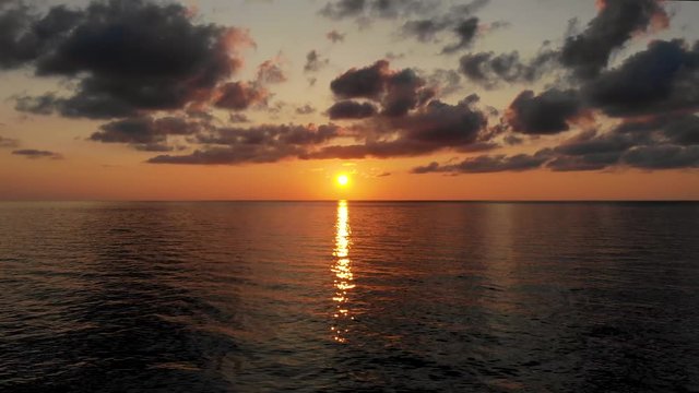 Flight over water to towards the fascinating landscape: sun setting in middle of sea and dark contrast clouds. Waves barely wave, sun path is reflected in sea surface. Taken by drone at sunset