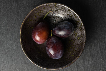 Group of three whole sweet purple plum on glaze bowl with water flatlay on grey stone