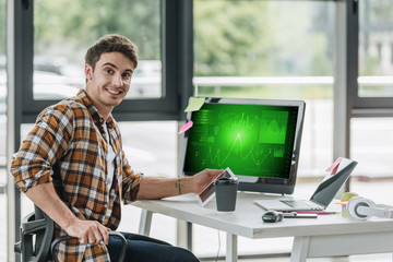 cheerful programmer smiling at camera while sitting near computer monitor with graphs and charts on...